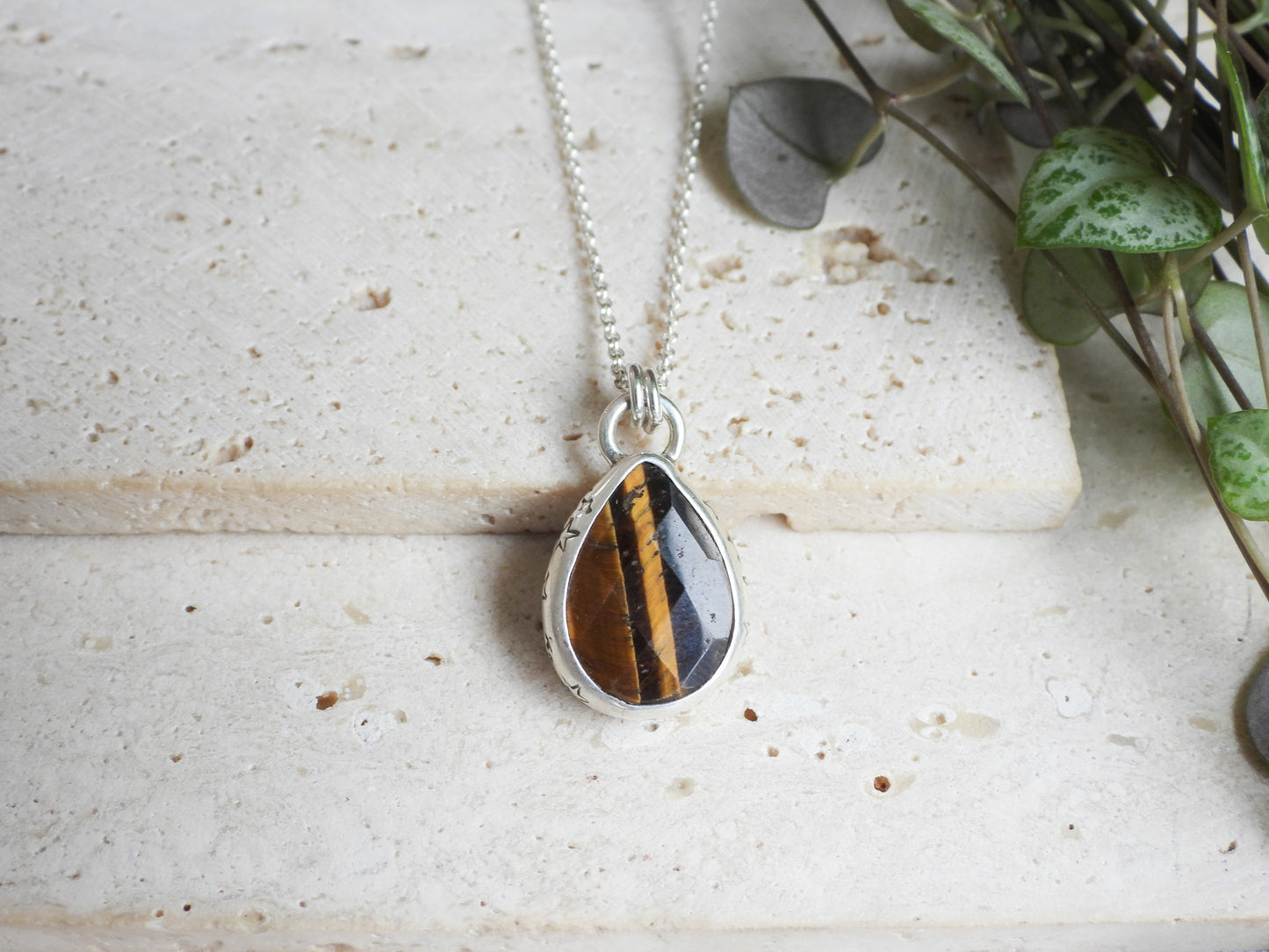 Teardrop-shaped pendant with tiger's eye stone on a silver chain, placed on a textured surface with greenery.