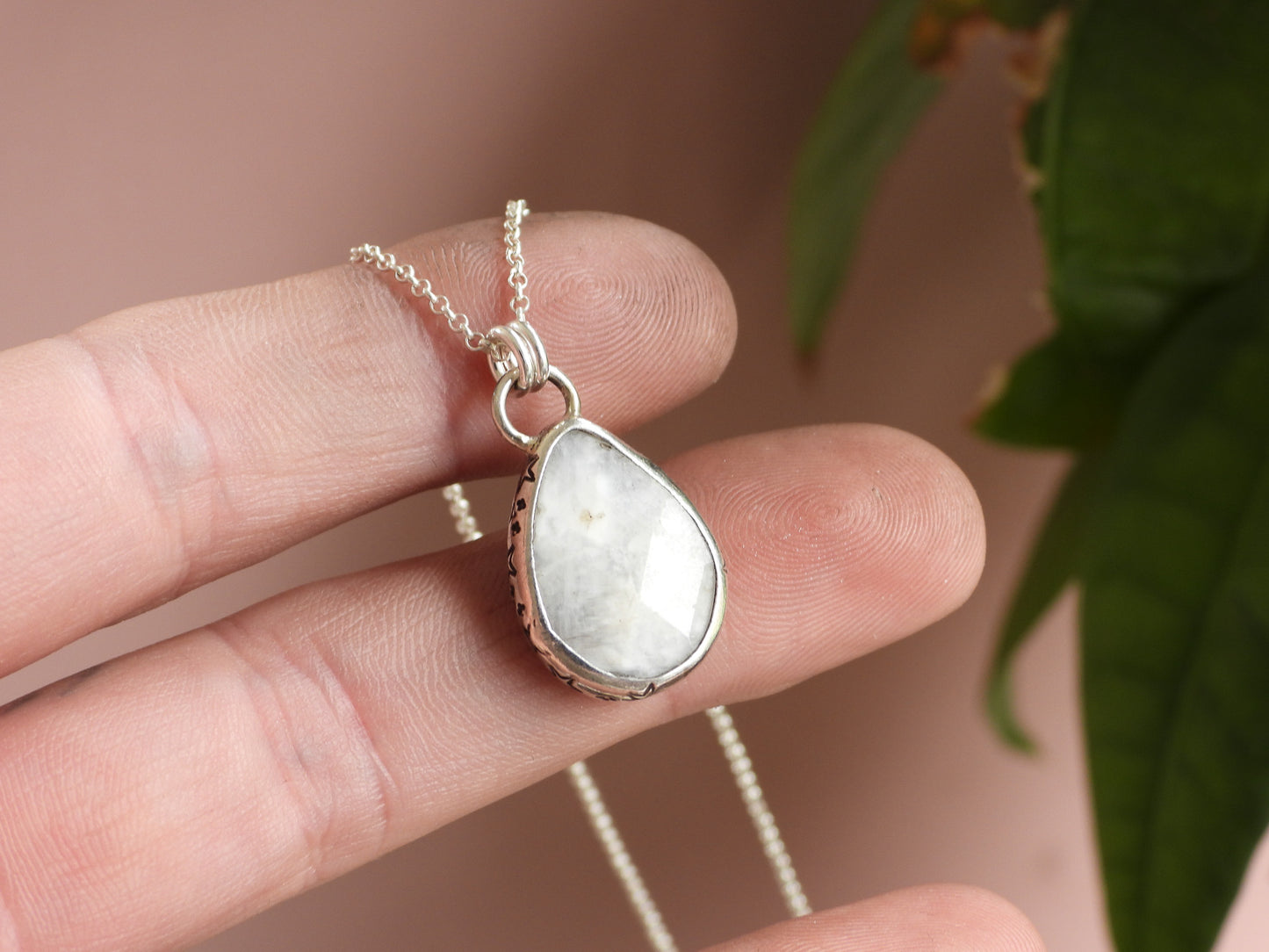 Teardrop-shaped moonstone pendant on a chain held between fingers with a blurred background