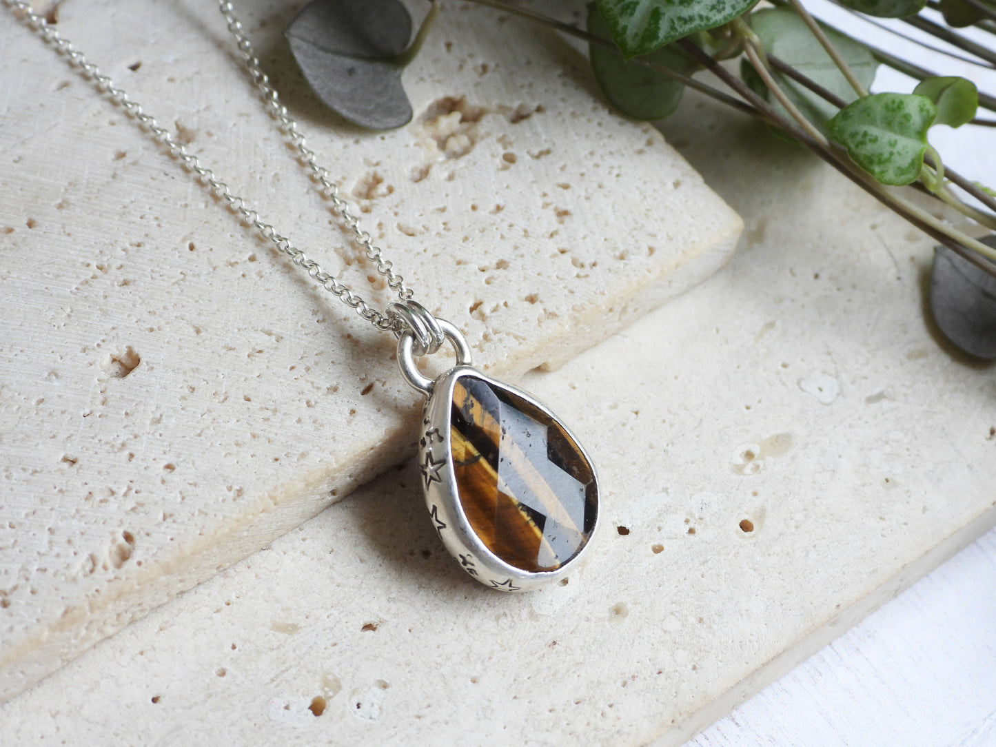 Teardrop-shaped tiger's eye pendant on a silver chain with a natural stone pattern, placed on a textured surface with greenery.