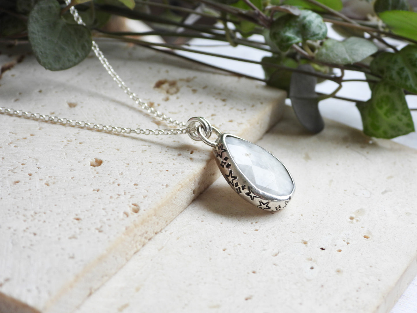 Silver moonstone necklace with a teardrop-shaped pendant on a light surface with green leaves in the background