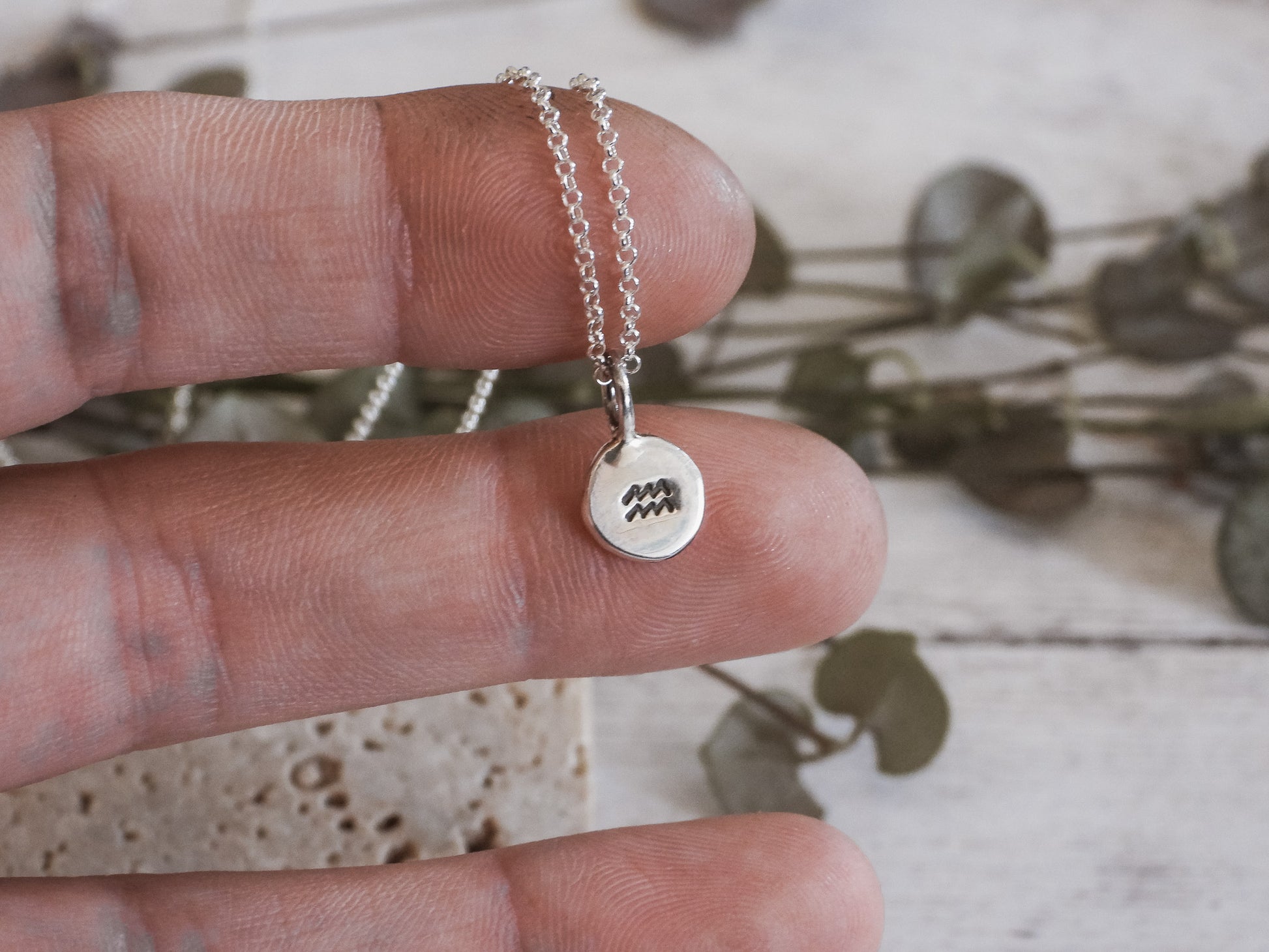Silver Aquarius necklace with a round pendant held between fingers on a light wooden surface.
