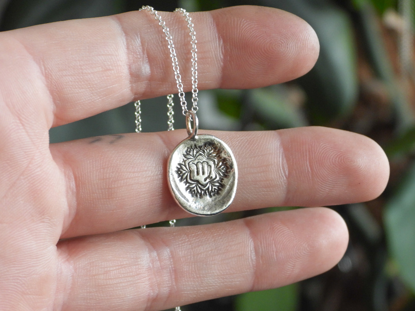 Silver virgo necklace with a round pendant held between fingers against a blurred green background