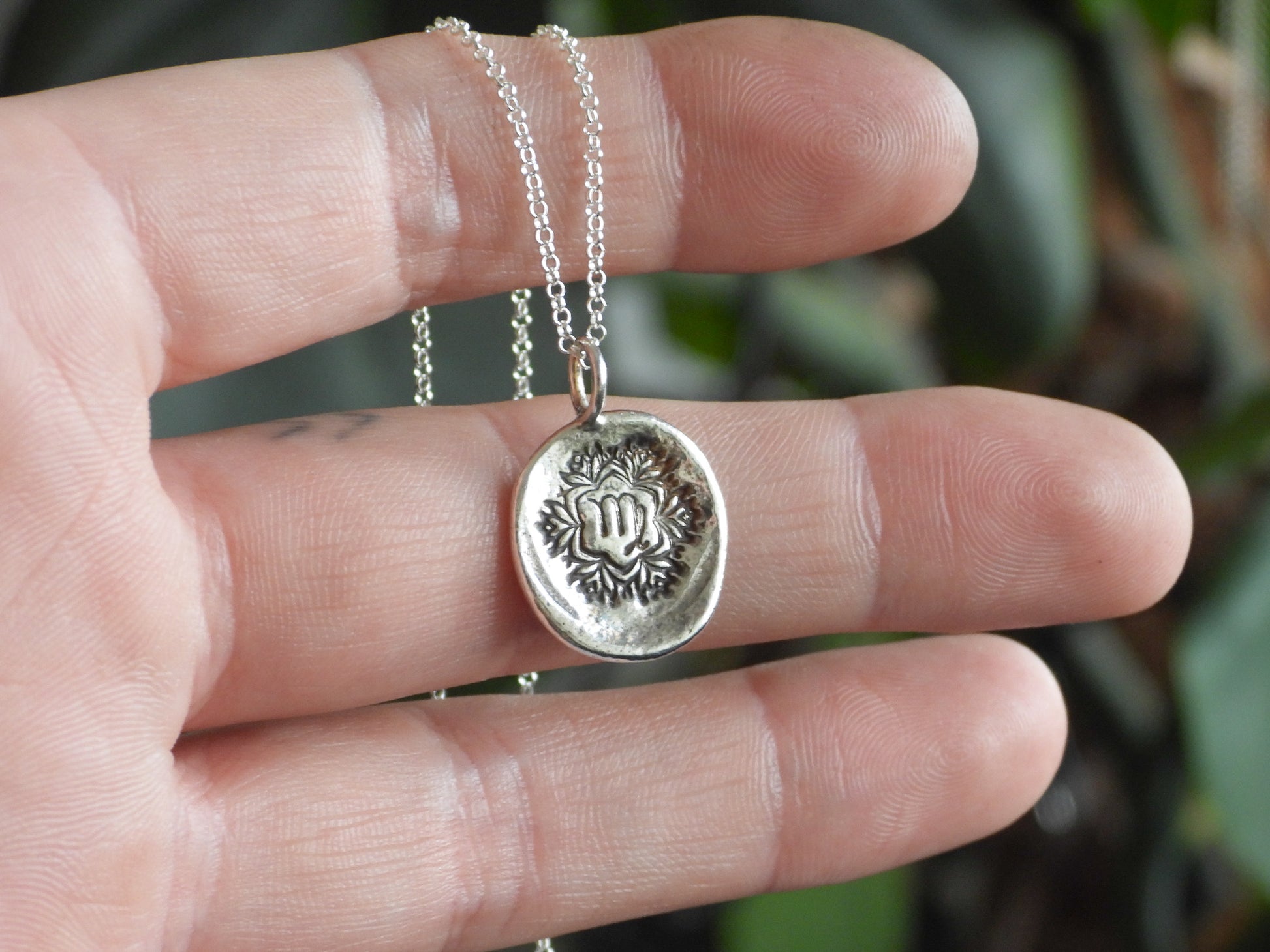 Silver virgo necklace with a round pendant held between fingers against a blurred green background