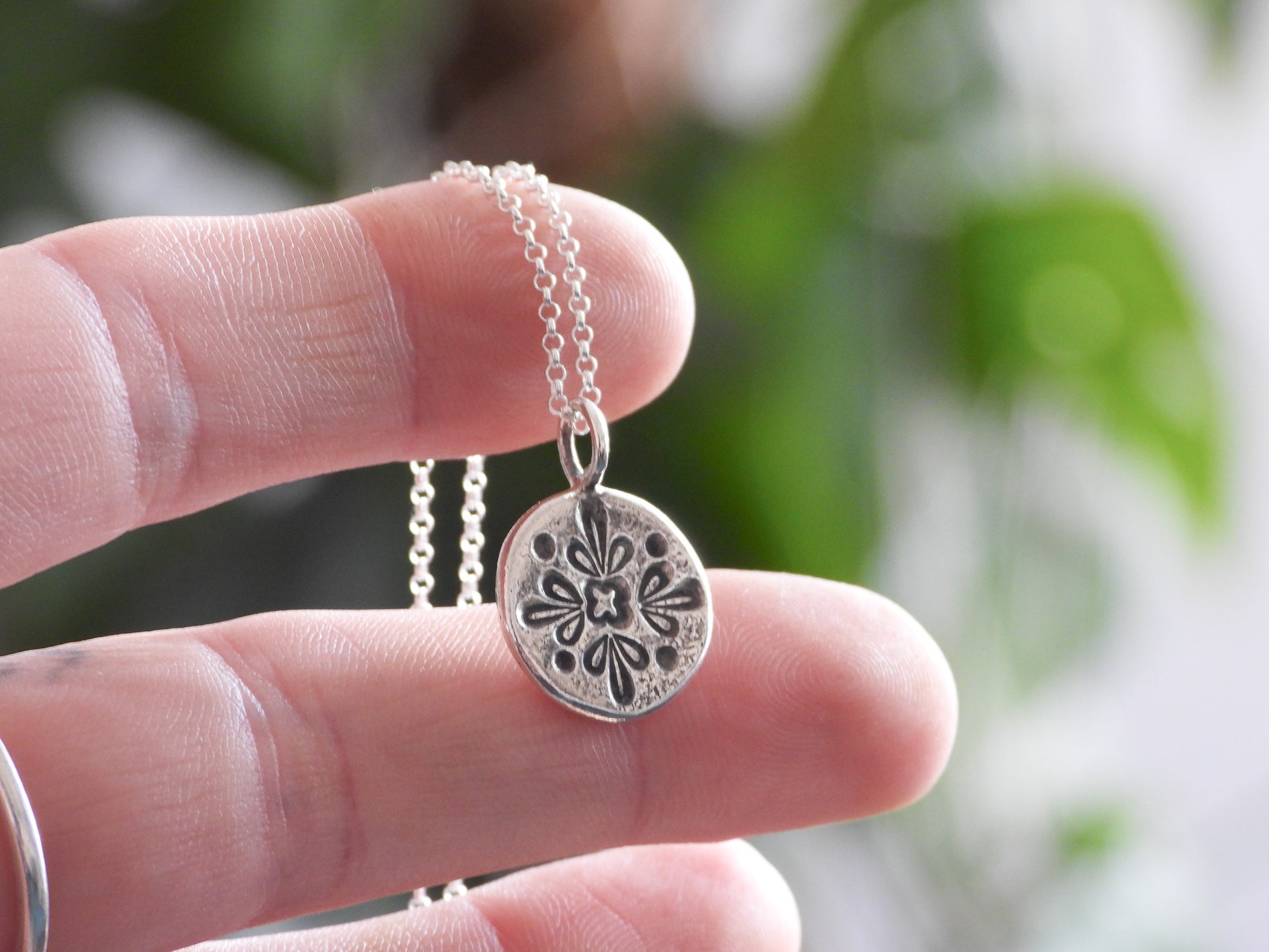 Silver mandala necklace with a floral pendant held between fingers against a blurred natural background