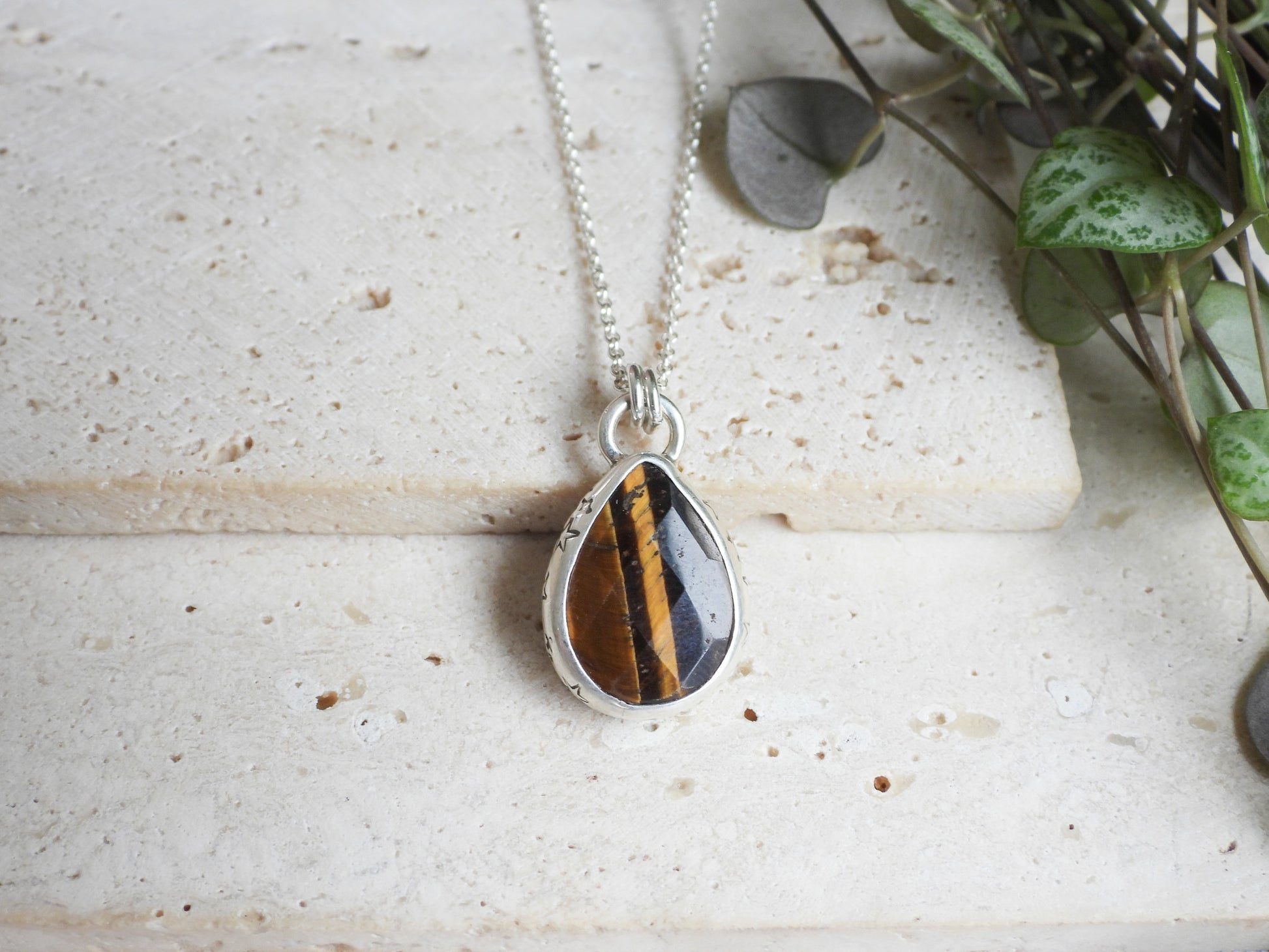 Teardrop-shaped pendant with tiger's eye stone on a silver chain, placed on a textured surface with greenery.