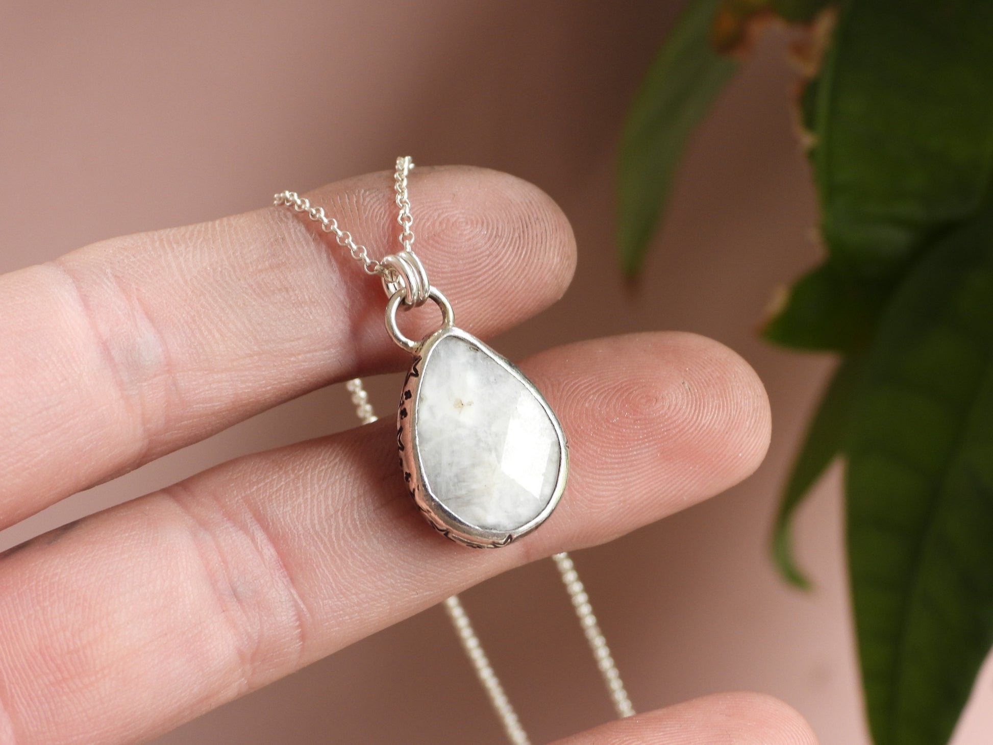 Teardrop-shaped moonstone pendant on a chain held between fingers with a blurred background