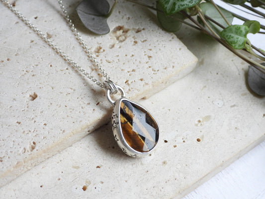 Teardrop-shaped tiger's eye pendant on a silver chain with a natural stone pattern, placed on a textured surface with greenery.