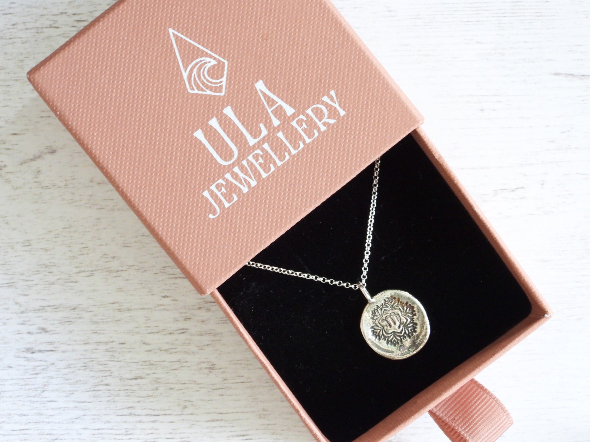 Silver necklace with a round pendant in a pink 'Ula Jewellery' box on a light wooden surface.