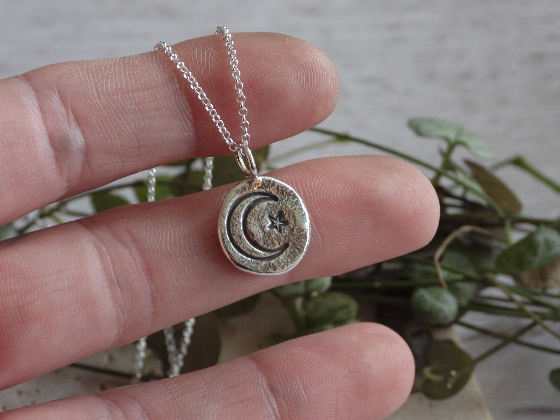 Silver necklace with a circular pendant held between fingers against a blurred natural background